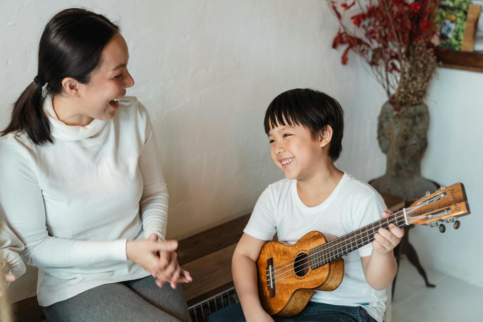 4elpapeldelamusicoterapiaeneltratamientodelautismo Niño étnico positivo sentado con un instrumento musical tradicional hawaiano y mirando hacia otro lado cerca de una madre contenta sentada con la boca abierta cerca de un jarrón con una rama decorativa