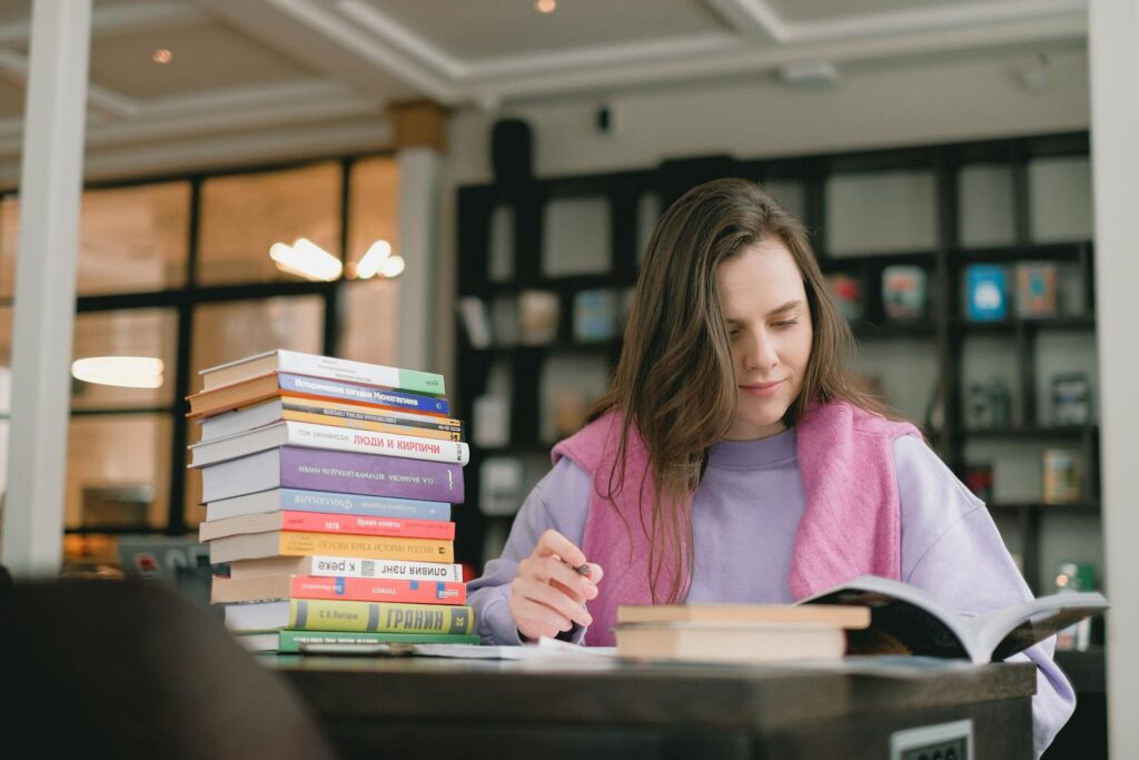 Female student in casual clothes sitting at table with books and writing notes while preparing for exam in university library